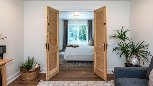 Ett sovrum med en stor säng, trädörrar, ett sängbord, en lampa, en krukväxt och utsikt över omgivningen.