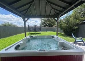 A hot tub under a covered patio with a grassy yard and a wooden fence in the background.