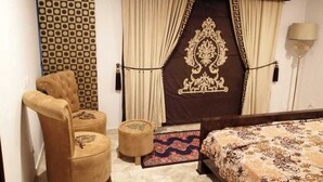 A hotel room with a bed, a chair, two ottomans, a patterned rug, and curtains with a decorative design. - Captivating 1-bed Apartment Fully Furnished (Lahore)