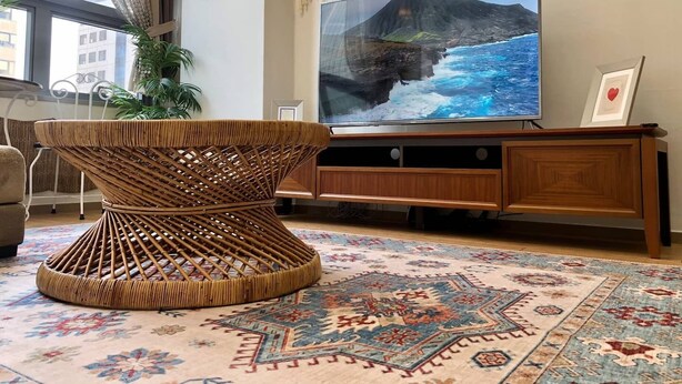 Ein Wohnzimmer mit einem großen Flachbildfernseher, einem gewebten Couchtisch, einem gemusterten Teppich und einer Topfpflanze.