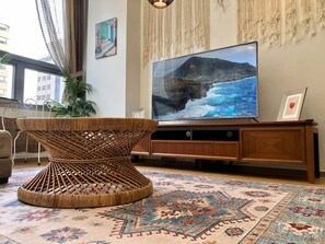 Uma sala de estar com uma televisão de tela plana grande, uma mesa de centro trançada, um tapete estampado e um vaso com planta.