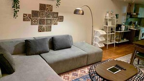 Ein Wohnzimmer mit grauem Sofa, Wandfliesen mit Muster, einer Stehlampe und einer Küchenzeile mit weißem Regalschrank.