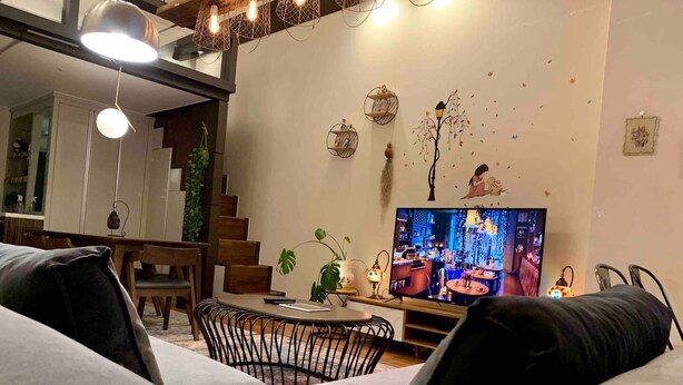 Ein modernes Wohnzimmer mit einer Couch, einem Esstisch und Stühlen. Ein Fernseher ist an der Wand montiert, und es gibt dekorative Wandbilder.