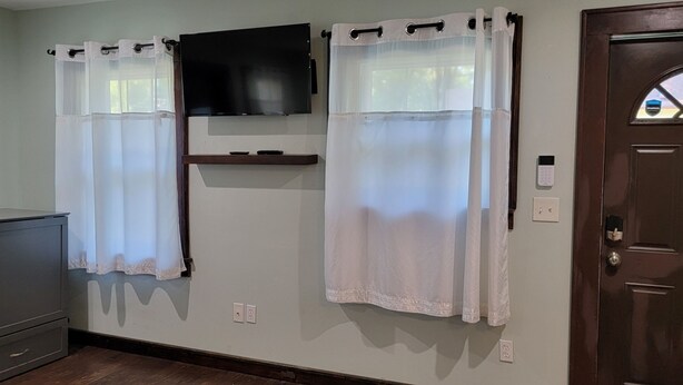 A room with a wooden door, a mounted flat-screen TV, and two windows with sheer curtains. - KC Tiny Studio (Kansas City)