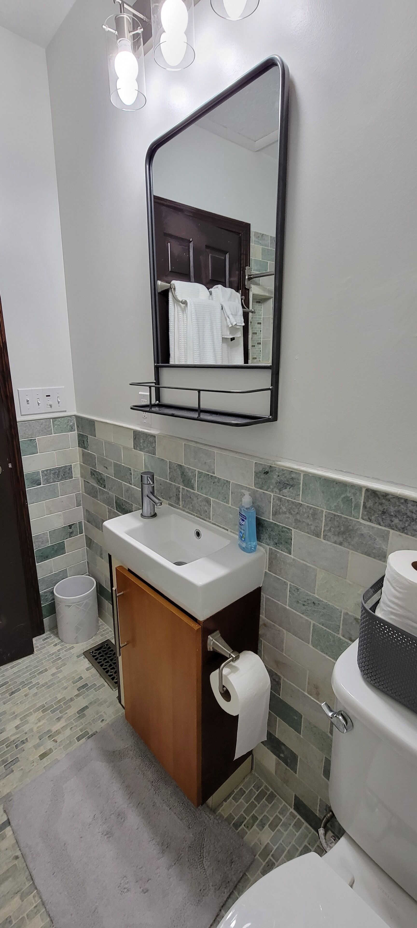 Une salle de bain avec un lavabo, un miroir et des toilettes blancs. Il y a un porte-serviettes au-dessus du lavabo et une poubelle à côté des toilettes.