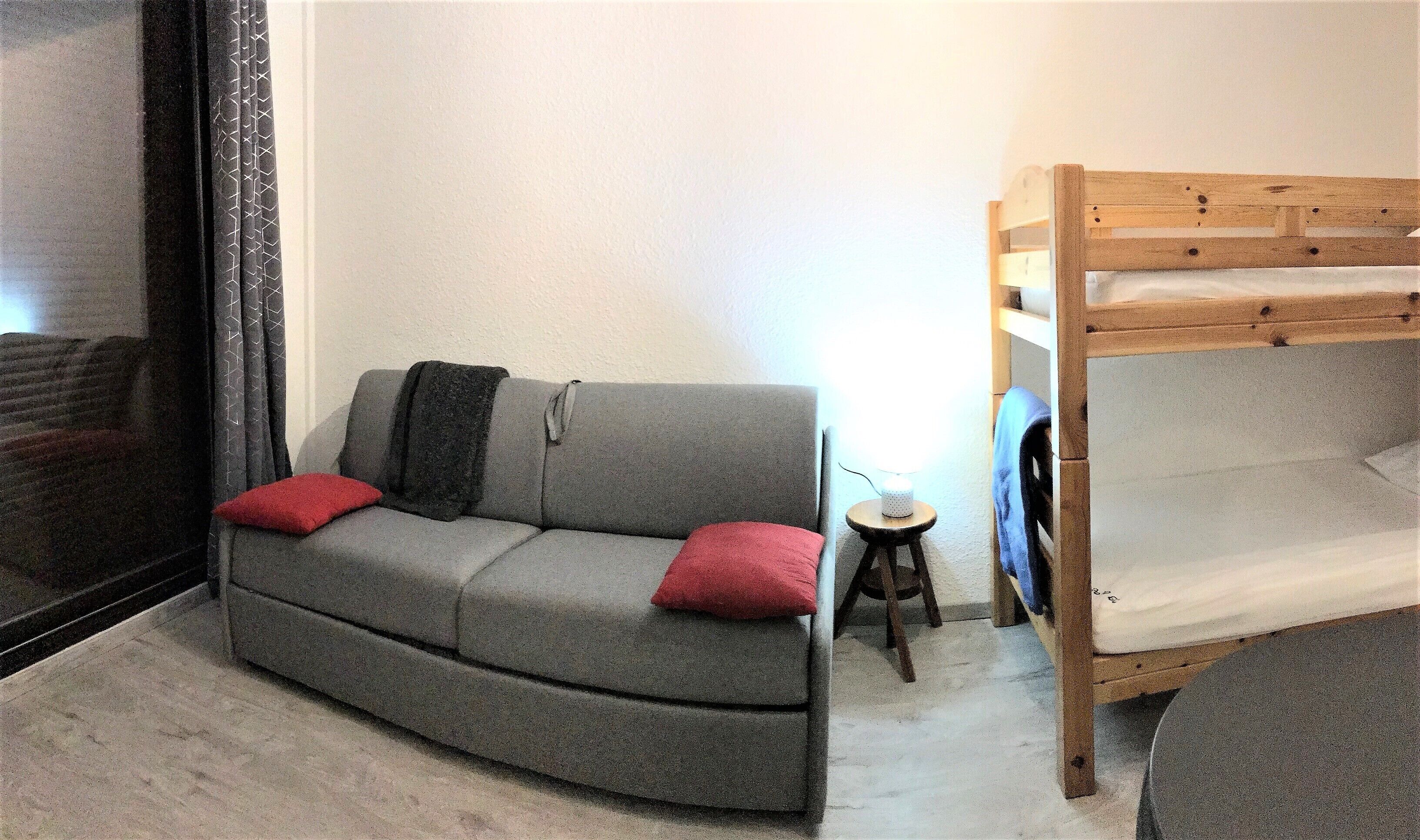 A small, compact room with a grey sofa, a wooden bunk bed, and a small stool.
