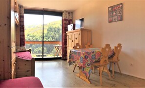 A room with a bed, a table with chairs, a TV on a wooden cabinet, and a view of a green landscape through a large window.