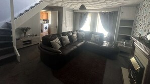 A living room with a dark leather sofa, a staircase, a kitchen, and a patterned wallpaper. - 3 Bedroom Miners Cottage - Wanlockhead D & G (Wanlockhead)
