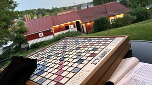 Un plateau de jeu de Scrabble avec des lettres et un cadre en bois, posé sur une table, avec en arrière-plan une maison au toit rouge et de la verdure.