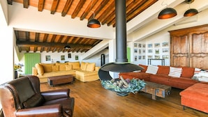 Un ampli saló amb un sostre alt, bigues de fusta, una llar de foc gran i negra, i diferents opcions de seients, com un butac armchair de cuir, un sofà modular groc i un sofà vermell.