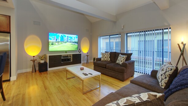 A modern living room with a flat-screen TV, a coffee table, a sofa, and a large window with blinds. - Houston Heights Lux 3 story-townhouse-Rice Military (Houston)