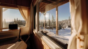 A cabin with a bed, a laptop on a table, and a view of a snowy forest through large windows.