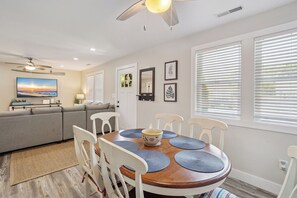 A dining area with a wooden table, white chairs, and blue placemats. There is a gray sofa and a TV in the background.