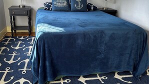 Quarto com colcha azul-marinho, duas almofadas iguais, tapete com padrão de âncora azul-marinho e branco, e duas mesas de cabeceira com luminárias.