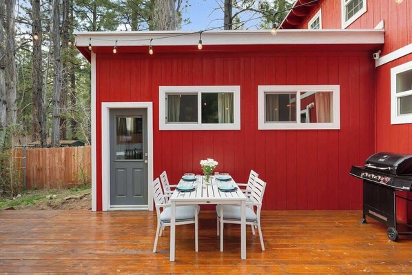 A red building with a white door and windows, a wooden deck, a barbecue grill, and a table with chairs.