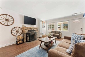 A living room with a stone fireplace, a television mounted on the wall, and a large window providing natural light.