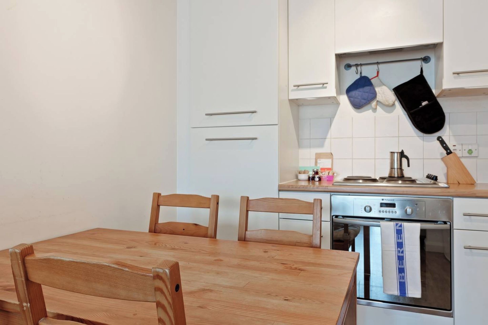 A compact kitchen with a wooden table and chairs, a built-in oven, and a hanging pot rack.