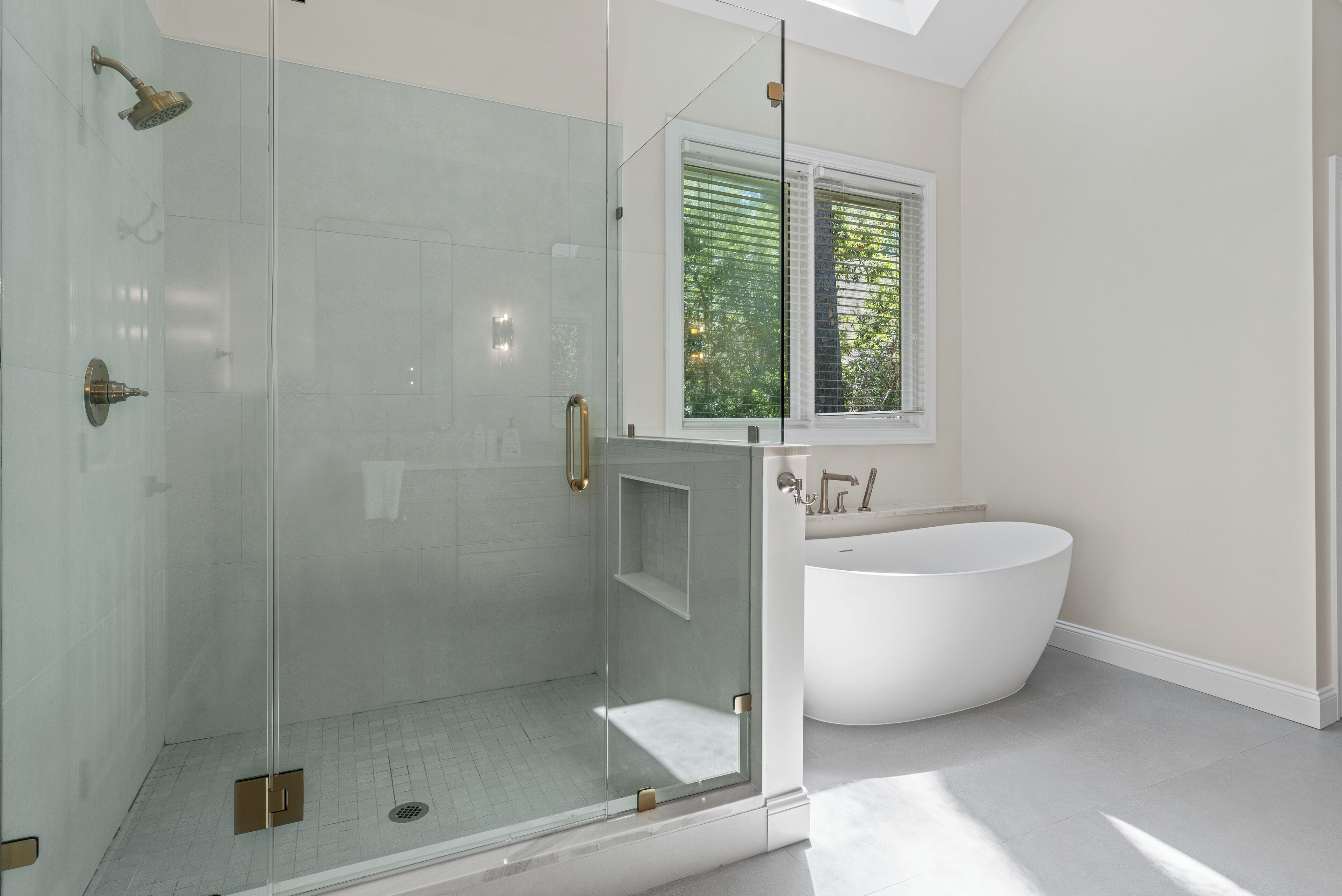 A modern bathroom with a glass-enclosed shower, a freestanding bathtub, and a window with blinds.