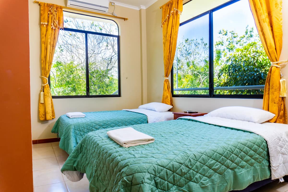 Quarto de hotel com duas camas, cada uma com colcha verde acolchoada e travesseiros brancos. Cortinas amarelas nas janelas, e o quarto oferece vista para o verde lá fora.