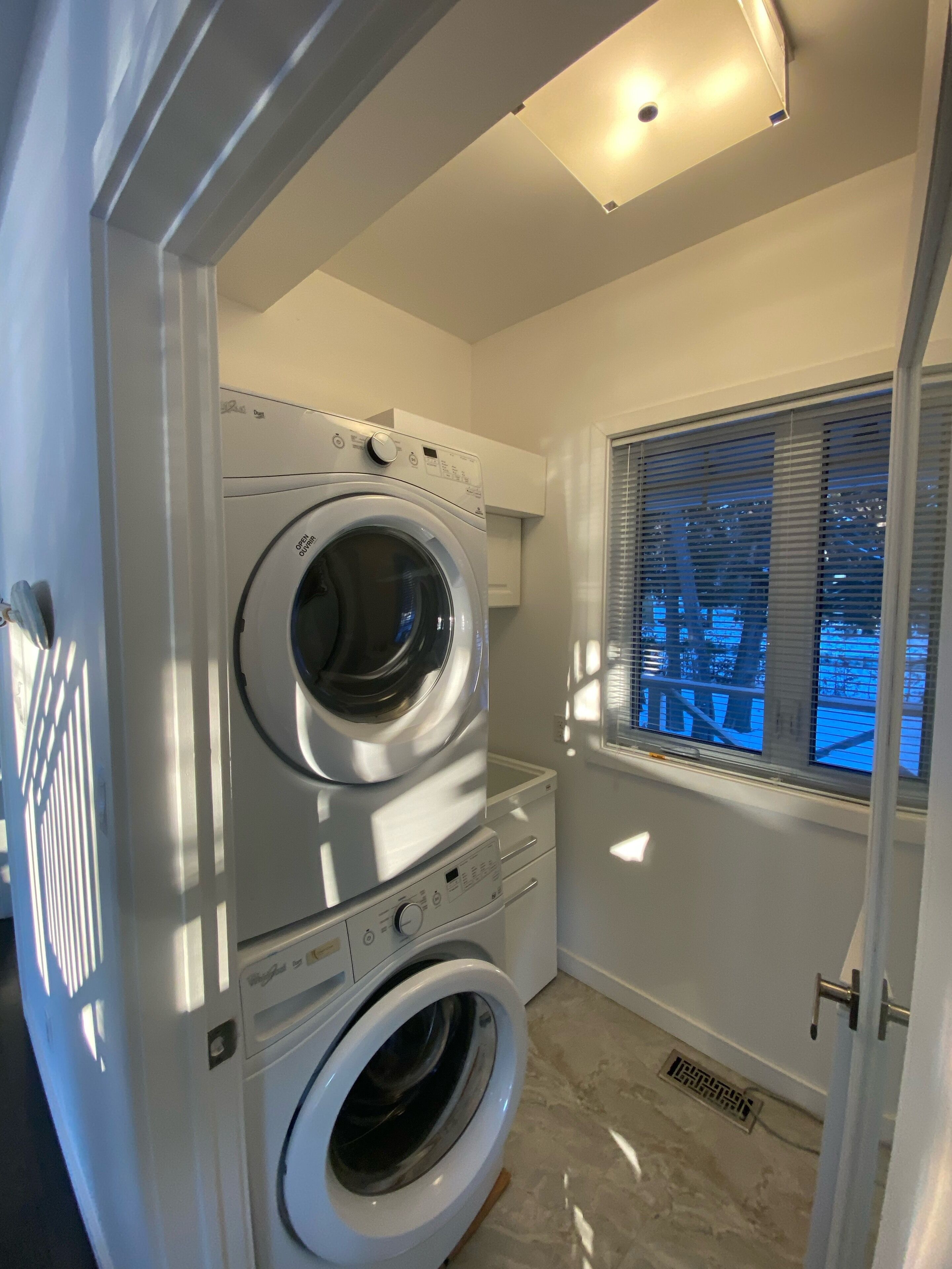 A laundry room with a front-loading washer and dryer, a window with blinds, and a vent on the floor.
