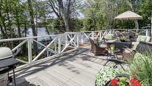 Een houten terras met buitenmeubilair, een barbecue en een witte reling met uitzicht op water.