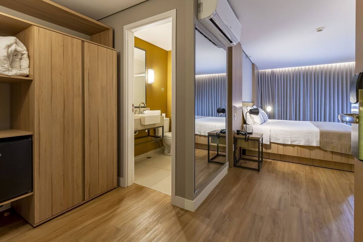 Una habitación de hotel con un armario de madera, un baño con lavamanos e inodoro, una cama con mesita de noche y un equipo de aire acondicionado.