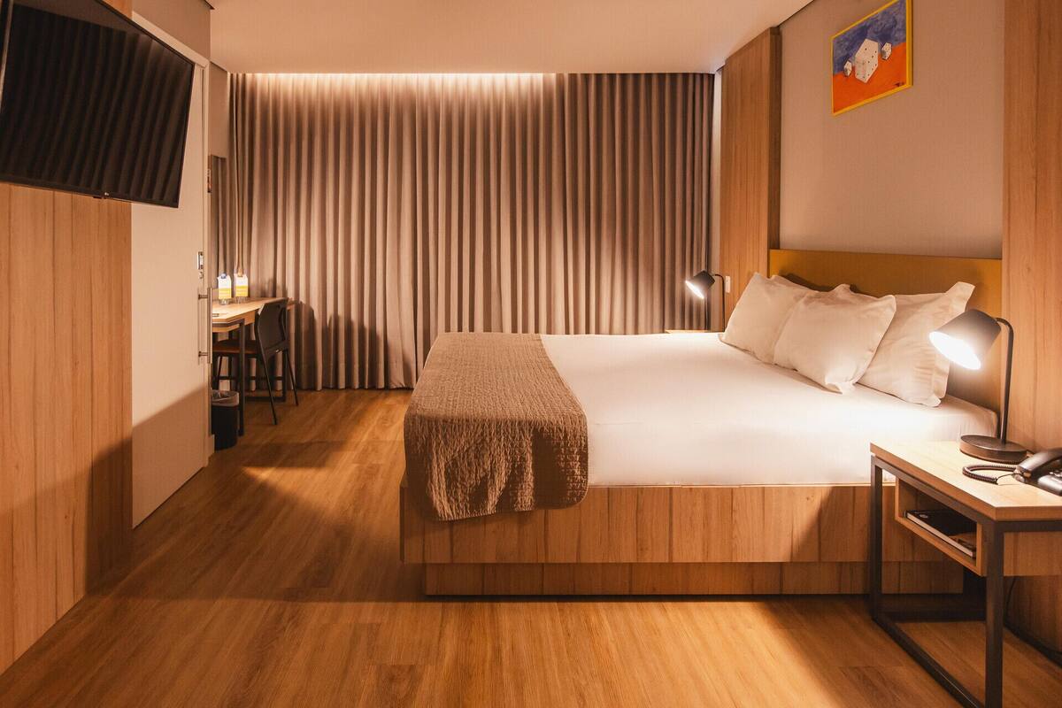 Una habitación de hotel con cama, mesita de noche, lámpara, escritorio, silla y cortinas.
