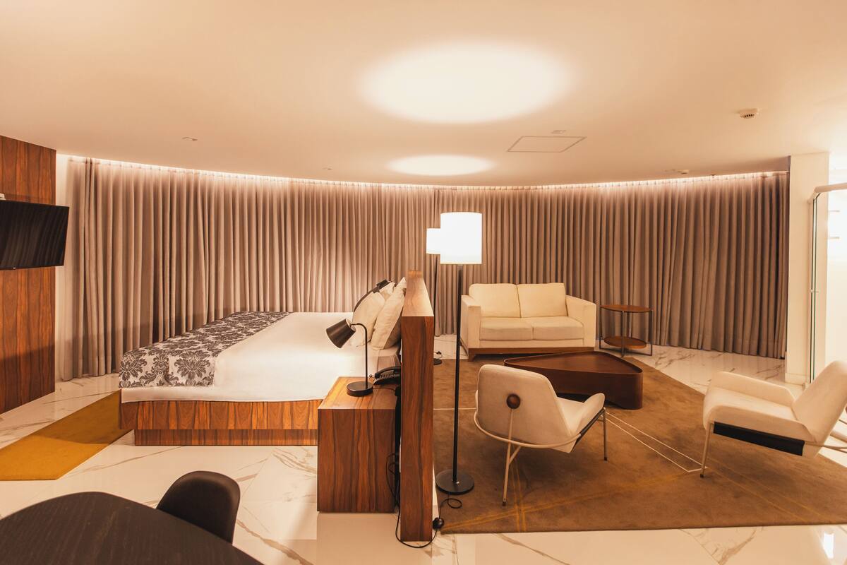 Una habitación de hotel moderna con una cama grande, una zona de estar con sofá y sillones, un escritorio y un televisor de pantalla plana montado en la pared.