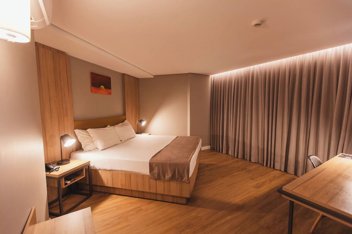 Habitación de hotel moderna con una cama de madera, mesitas de noche, un escritorio y un ventanal con cortinas.