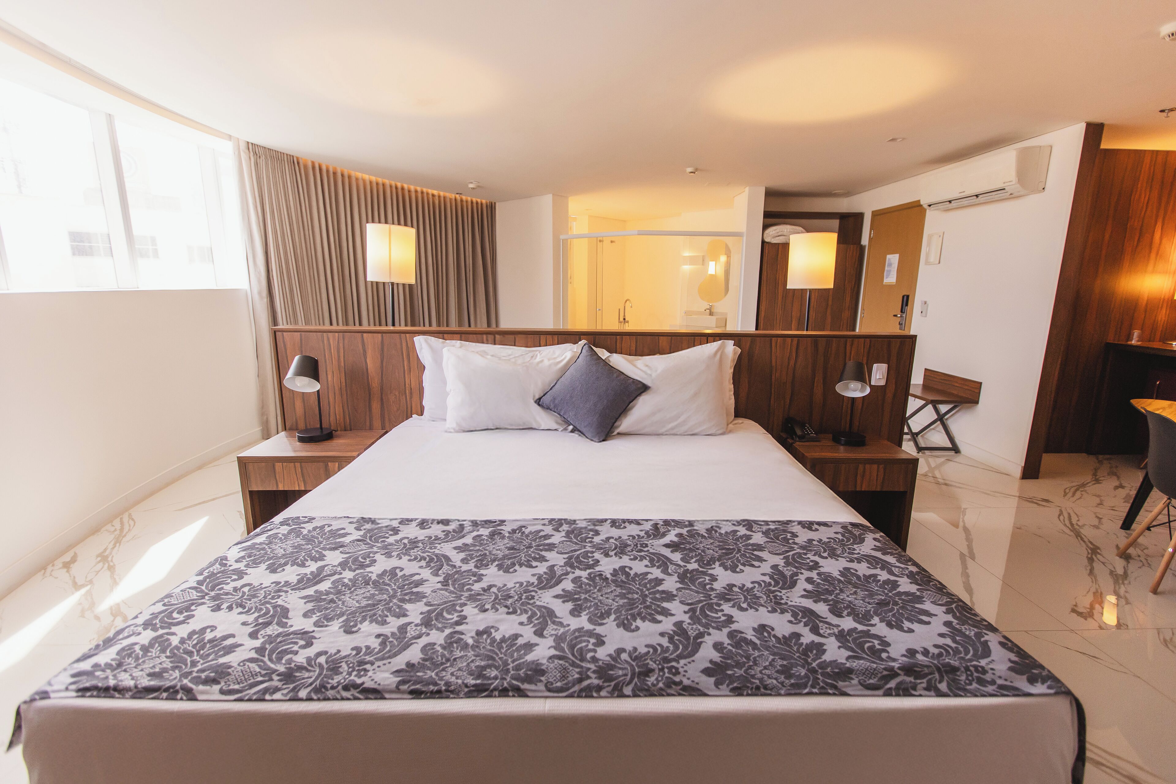 Habitación de hotel con una cama grande, un cabecero de madera, una mesita de noche con lámpara, un escritorio con silla, un espejo y un baño visible a través de una puerta de cristal.