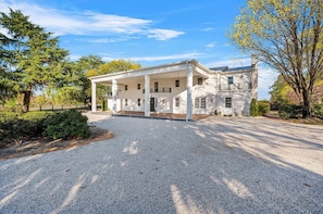 A two-story white building with a covered porch, a gravel driveway, and surrounding trees.