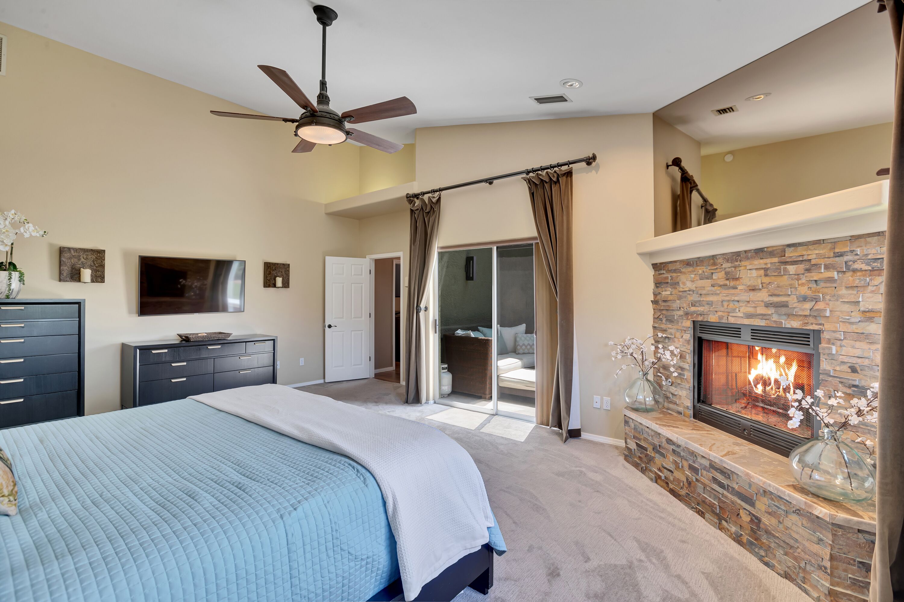 A bedroom with a large bed, a fireplace, a ceiling fan, and a sliding glass door leading to a balcony.