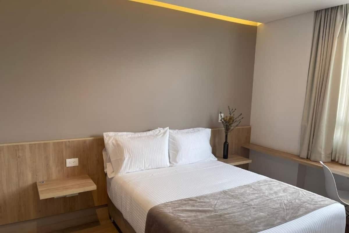 A hotel room with a bed, bedside tables, a nightstand, a vase with flowers, and a window with curtains.