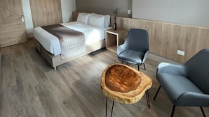 Quarto de hotel moderno com uma cama, duas cadeiras, uma pequena mesa de madeira e uma mesinha de cabeceira.