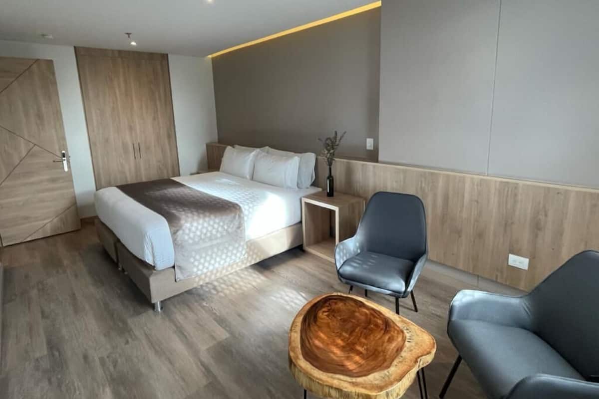 A modern hotel room with a bed, two chairs, a small wooden table, and a nightstand.