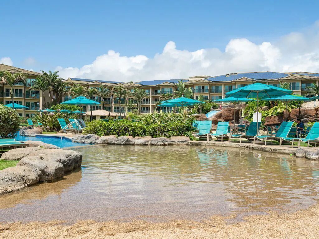 Un espace piscine aménagé dans un complexe hôtelier, avec des chaises longues et des parasols, entouré de palmiers et bordé d’un bâtiment doté de nombreux balcons.