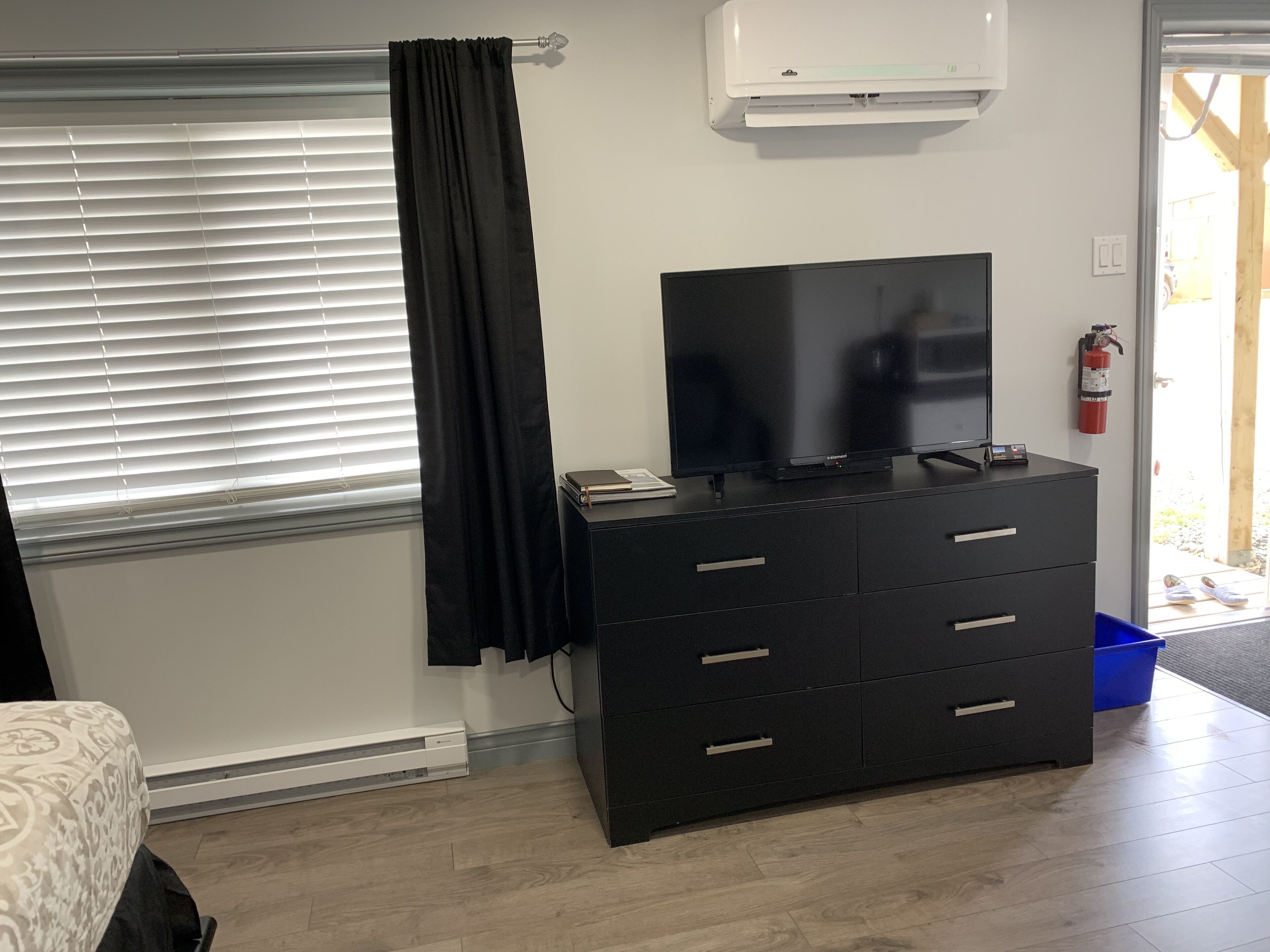 A room with a large flat-screen TV on a black dresser, a window with blinds, a white air conditioning unit, and a fire extinguisher mounted on the wall.