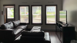 A living room with a leather sofa, a coffee table, and a TV on a stand. There are three windows with blinds, and a fire extinguisher is mounted on the wall. - La Digue Suites  (Cheticamp)