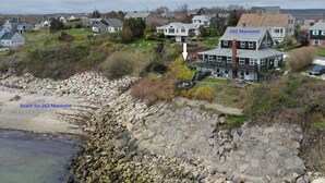 Une propriété située sur la côte, avec un escalier en pierre menant à une plage, une grande maison portant le numéro « 262 Manomet », et une vue sur le quartier environnant.