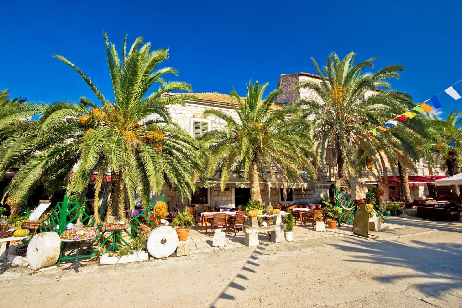 A coastal outdoor seating area with palm trees, stone buildings, and a clear blue sky.