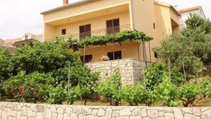 A two-story house with a stone wall in front, a balcony with a metal railing, and a garden with green shrubs and red flowers.