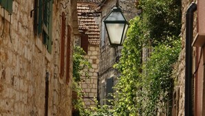 A narrow cobblestone street flanked by historic stone buildings with green shutters and a hanging street lamp.