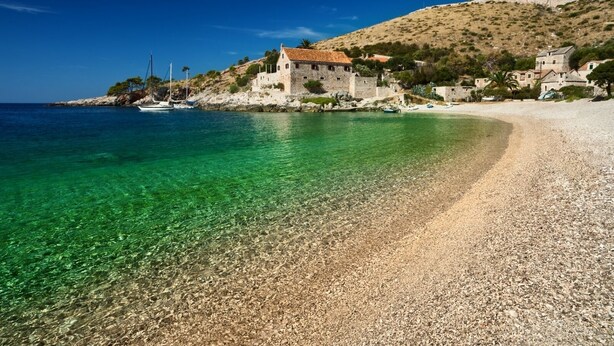 A coastal scene with a pebble beach, clear turquoise water, and a stone building with a red-tiled roof.