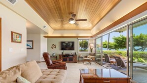 A spacious living room with a large sofa, a coffee table, and a television. There is a ceiling fan, a fireplace, and a view of the outdoors through large windows. - Puunoa Beach Estates- Part OF Hyatt (LAHAINA)