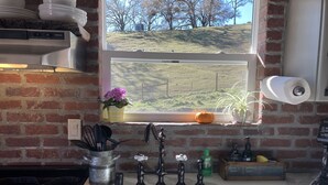 A kitchen with a white sink, a window with a view of a grassy hill, a brick wall, and a shelf with kitchen utensils. - Romantic Private Getaway!  Hot tub, Fireplace and Views!  (Hopland)