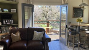 A cozy living room with a brown leather sofa, a bookshelf, a dining area with a table and chairs, and a large sliding glass door leading to a balcony with a view of trees.