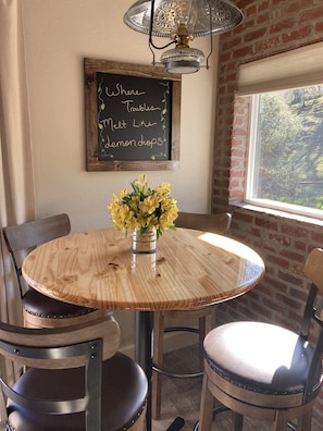 Une table ronde en bois, sur laquelle est posé un vase de fleurs jaunes, deux chaises, un tableau noir avec des inscriptions et un mur de briques avec une fenêtre.