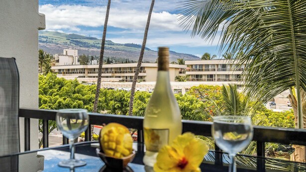 Un balcón con una mesa dispuesta para dos personas, que incluye una botella y dos copas de vino, y vistas a un edificio y follaje al fondo.