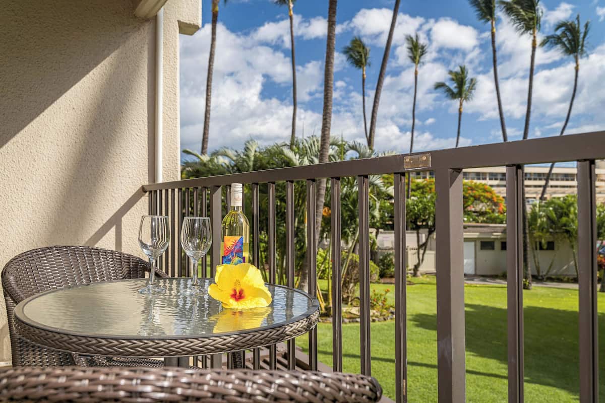 A round glass table with wicker chairs on a balcony, overlooking a garden with palm trees and a clear sky.