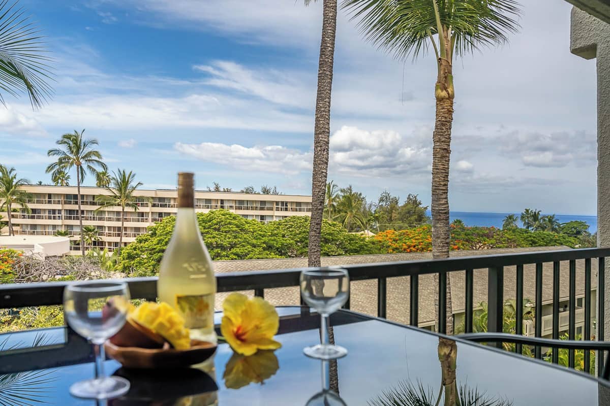 A table with a bottle of wine, a glass, and a flower, overlooking a hotel with palm trees and a view of the ocean.